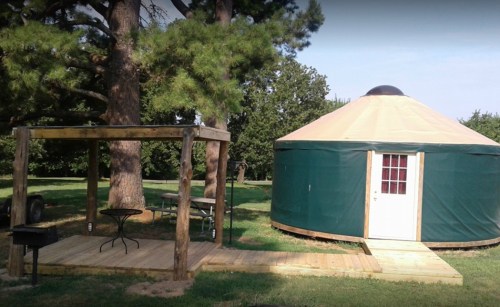 A green yurt with a wooden deck and table, surrounded by trees in a grassy area.