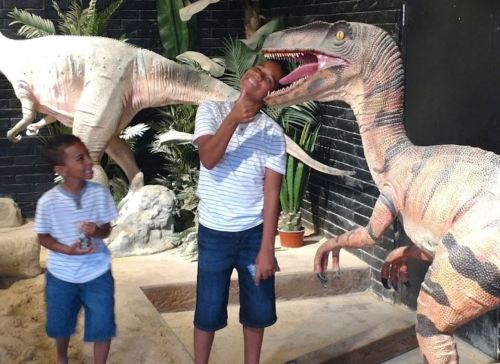 Two boys pose playfully with a life-sized dinosaur model in a museum exhibit. One boy mimics a dinosaur bite.
