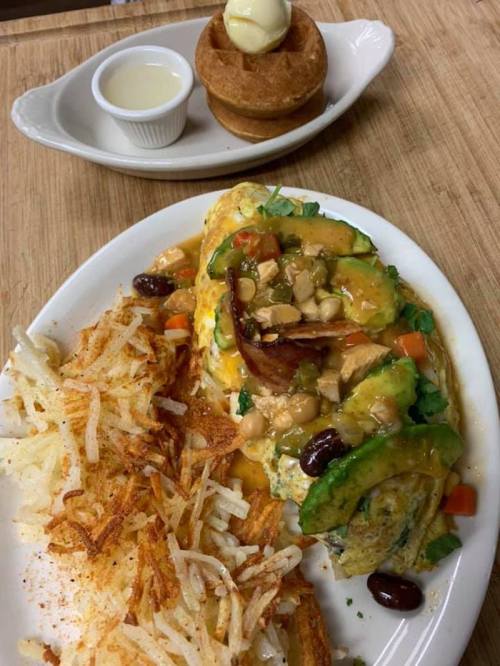 A plate with a colorful omelette topped with beans and avocado, served with crispy hash browns and a waffle with butter.