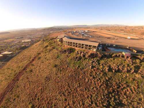 Aerial view of a hotel on a hillside, surrounded by dry landscape and distant mountains under a clear sky.