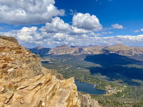 A panoramic view of mountains and a lake under a blue sky with fluffy clouds, seen from a rocky cliff.