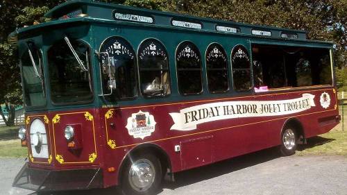 A maroon trolley bus with decorative windows, labeled "Friday Harbor Jolly Trolley," parked in a green area.