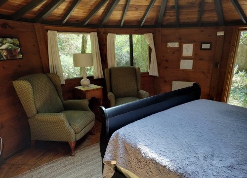 Cozy wooden interior of a cabin featuring a bed, two armchairs, a lamp, and large windows with natural light.