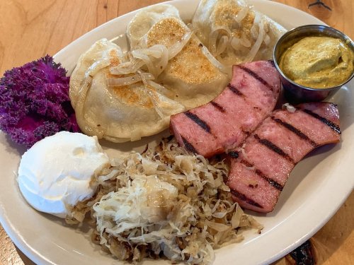 A plate featuring pierogi, grilled sausage, sauerkraut, sour cream, and a small bowl of mustard, garnished with purple cabbage.