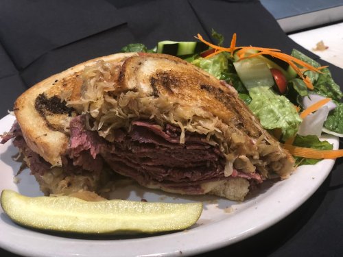 A stacked sandwich with corned beef and sauerkraut, served with a side salad and a pickle slice.