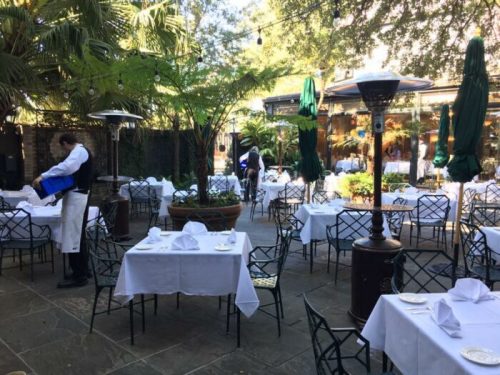 Outdoor dining area with empty tables set for guests, surrounded by greenery and a waiter preparing the space.