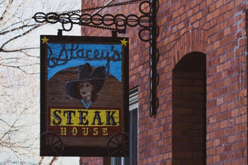 A wooden sign for "Marcus's Steak House" featuring a cowboy hat and a portrait, hanging from a brick building.
