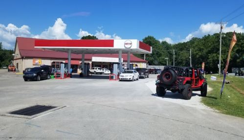 A gas station with a red roof, several cars parked, and a bright blue sky with clouds in the background.