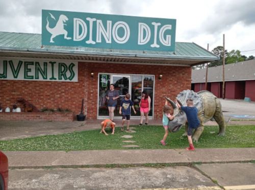 A group of children play outside a souvenir shop called "Dino Dig," featuring a dinosaur statue on the lawn.
