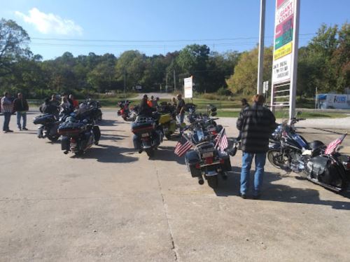 A group of motorcyclists gather at a gas station, with several bikes displaying American flags in a scenic setting.
