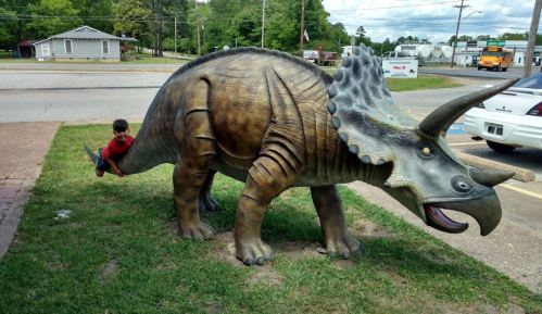 A child playfully interacts with a large dinosaur statue in a grassy area near a road.