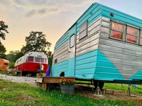 A vintage turquoise and silver trailer parked beside a red Airstream, set against a sunset sky.