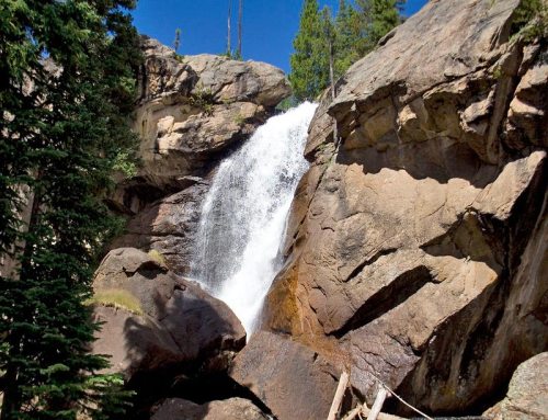 A waterfall cascading down rocky cliffs, surrounded by lush green trees under a clear blue sky.