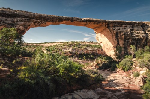 Natural stone arch spanning a canyon, surrounded by lush greenery and a clear blue sky.