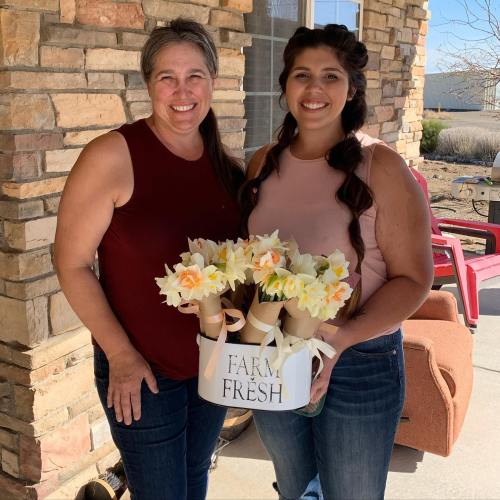 Two women smile while holding bouquets of flowers in a rustic outdoor setting. A "Farm Fresh" sign is visible.