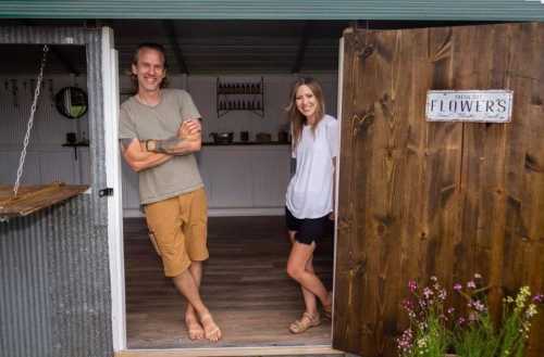 A man and woman stand at the entrance of a rustic shop, smiling, with flowers and greenery around them.