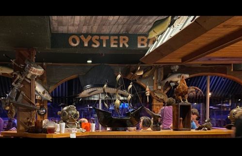 A lively restaurant interior featuring nautical decor, including fish, a ship model, and diners enjoying their meals.