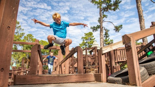 A child jumps over a wooden structure at a playground, while another child runs in the background under a blue sky.