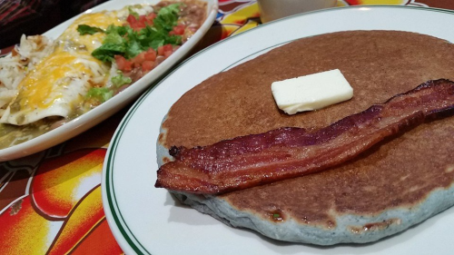 A plate featuring a large pancake with a slice of butter and crispy bacon, alongside a dish of enchiladas topped with cheese and vegetables.