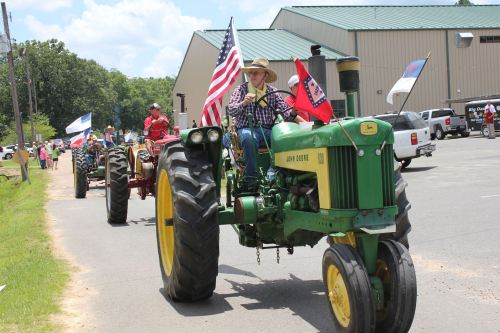A parade featuring tractors, with participants waving flags, driving down a rural road on a sunny day.