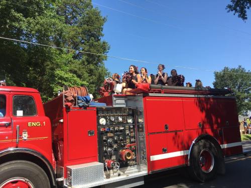 A red fire truck with people in colorful outfits sitting on top, enjoying a sunny day during a parade.