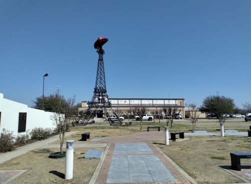 A small replica of the Eiffel Tower topped with a red hat, set in a grassy park with a clear blue sky.