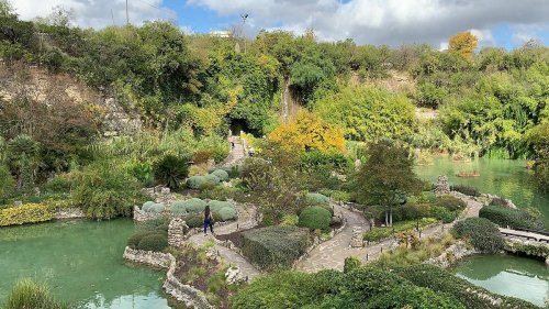 A serene garden with winding paths, lush greenery, and a tranquil pond under a partly cloudy sky.