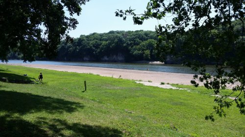 A serene riverbank scene with green grass, trees, and a sandy beach, featuring a few people enjoying the water.