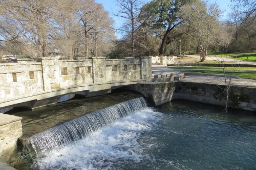 A stone bridge over a small waterfall, surrounded by trees and a peaceful park setting.