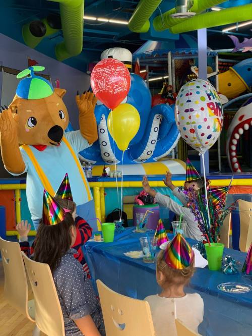 A costumed bear mascot greets children at a colorful birthday party with balloons and festive decorations.