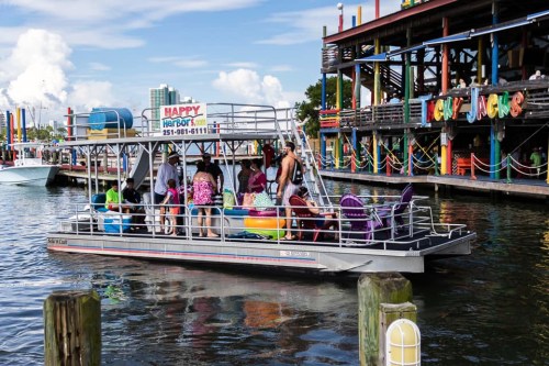 A party boat with colorful seating is docked near a lively waterfront restaurant on a sunny day.