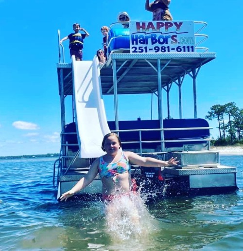 A girl splashes in the water while others play on a boat with a slide, enjoying a sunny day at the beach.