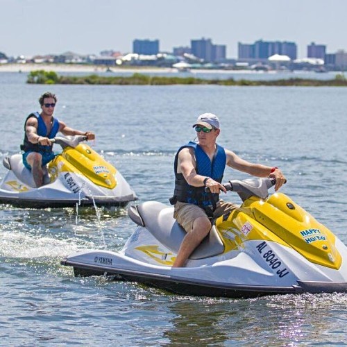 Two people riding yellow jet skis on a calm waterway with a city skyline in the background.