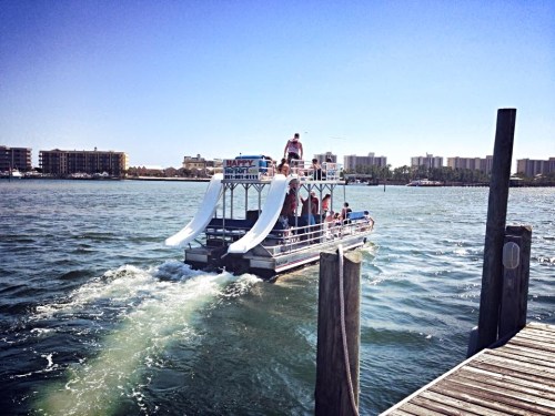 A party boat with water slides cruising on a sunny day, with people enjoying the ride in a scenic waterfront area.
