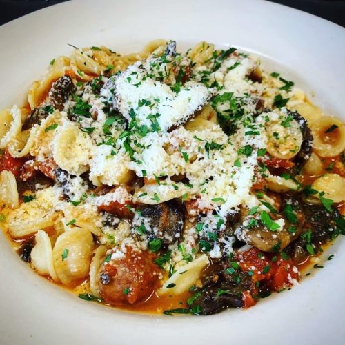 A bowl of orecchiette pasta topped with sausage, tomatoes, herbs, and grated cheese.