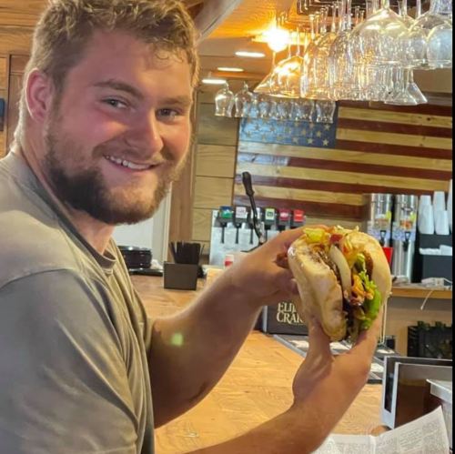 A smiling man holds a large sandwich in a restaurant with an American flag in the background.