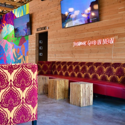 Colorful interior of a cafe with vibrant wall art, plush seating, wooden tables, and neon sign saying "You look good in neon."