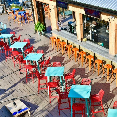 Colorful outdoor seating area with turquoise and red tables and chairs, string lights, and a bar in the background.