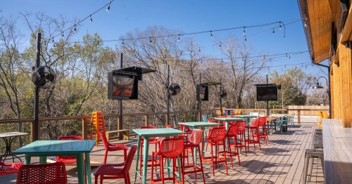 Outdoor patio with colorful tables and chairs, string lights, and TVs, surrounded by trees on a sunny day.