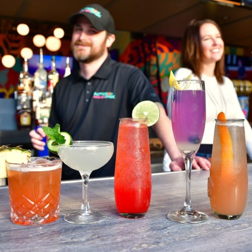 A bartender stands behind a bar with five colorful cocktails, smiling with a woman in the background.