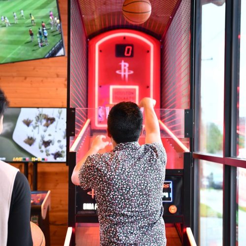 A person in a floral shirt shoots a basketball at an arcade game, with sports screens in the background.