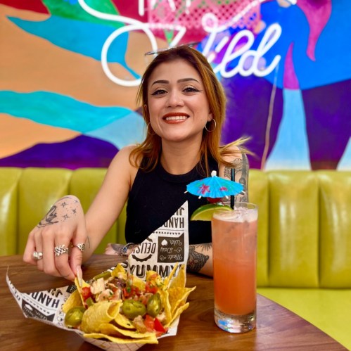 A smiling woman with tattoos enjoys nachos and a colorful drink in a vibrant restaurant setting.