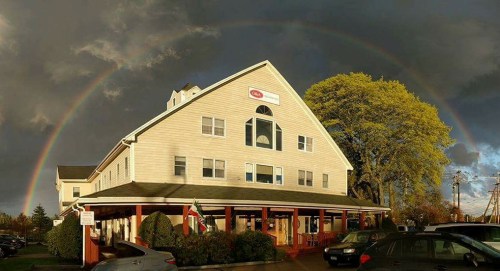 A large building with a peaked roof under a dramatic sky, featuring a rainbow and surrounded by greenery and parked cars.