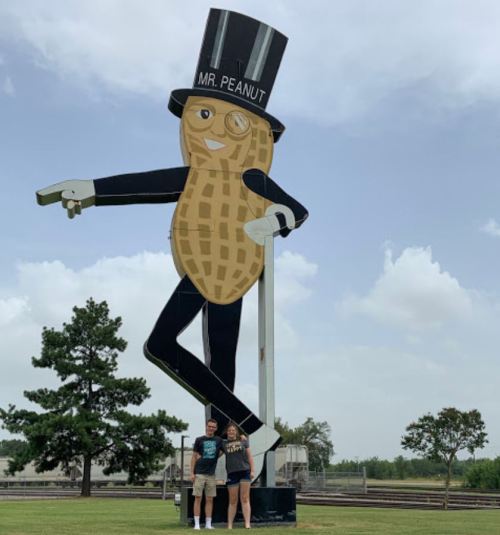 A couple stands in front of a large, cartoonish Mr. Peanut statue wearing a top hat and monocle, under a cloudy sky.