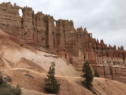 Rocky cliffs with unique formations and a natural arch, set against a cloudy sky. A dirt path winds through the landscape.
