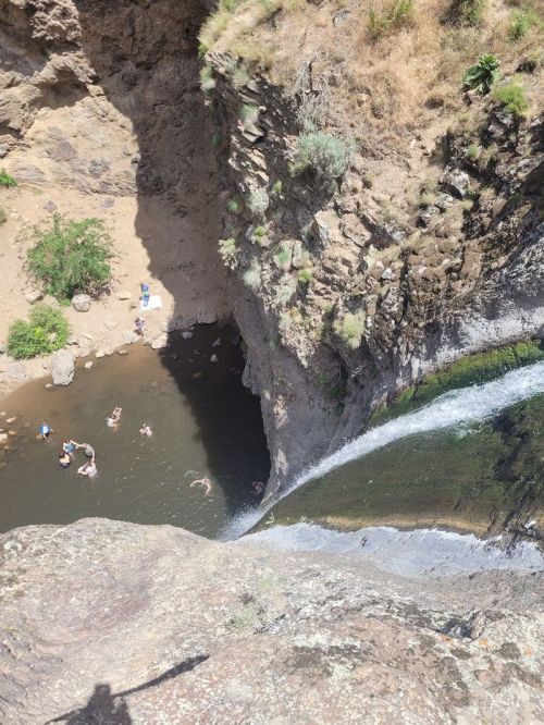 Aerial view of a waterfall cascading into a pool, with people swimming and relaxing nearby on a sunny day.
