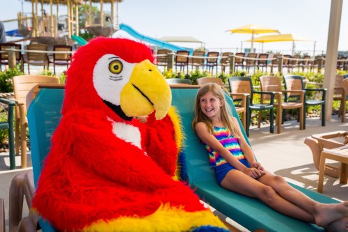 A girl smiles next to a large parrot mascot on a lounge chair by a poolside. Bright, sunny day with water slides in the background.
