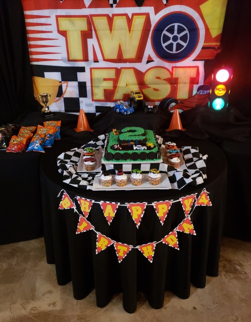 A birthday table with a race car theme, featuring a cake, cupcakes, snacks, and decorations.