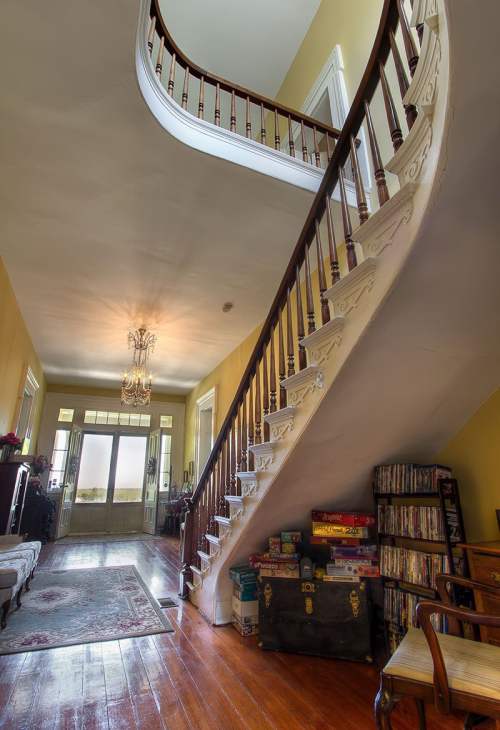 A spacious foyer with a curved staircase, chandelier, and vintage furniture, leading to large windows and a view outside.