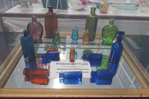 A display of colorful antique poison bottles in various shapes and sizes, labeled with the collector's name.